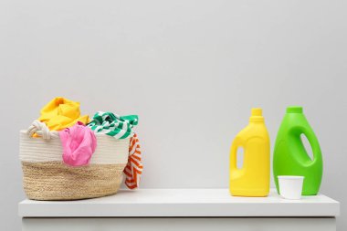 Çamaşır sepetinde giysi ve deterjan var. Dolabın yanındaki beyaz duvarda.