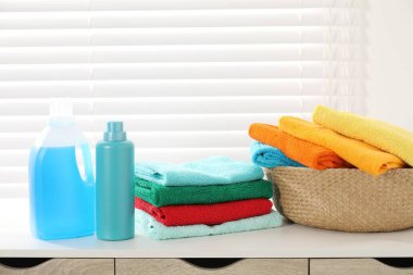 Çamaşır sepetinde havlular ve bir şişe deterjan var. Kapalı pencerenin yanındaki beyaz masada.