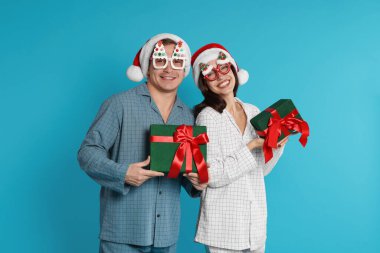Noel Baba şapkalı ve pijamalı mutlu çift açık mavi arka planda Noel hediyeleri
