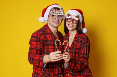 Noel Baba şapkalı ve pijamalı mutlu çift turuncu arka planda şeker kamışlarıyla kalp yapıyorlar. Noel atmosferi