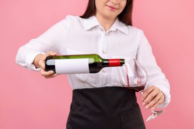 Profesyonel şarap garsonu pembe arka planda kırmızı şarabı bardağa dolduruyor.