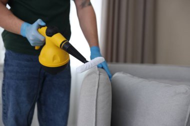 Haşere kontrol işçisi kanepeyi temizliyor. Kapalı alanda buhar temizleyicisi var.