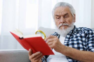 Üst düzey bir adam kanepede kitap okurken büyüteç kullanıyor.