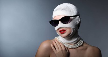 Gri arkaplandaki estetik ameliyattan sonra yüzü tıbbi bandaja sarılı bir kadın.