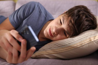 Genç çocuk evdeki kanepede akıllı telefon kullanıyor.