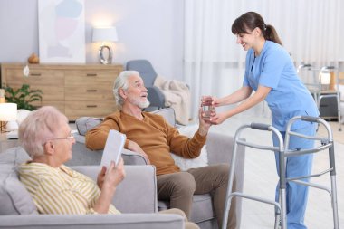 Huzurevinde kitap okuyan yaşlı erkek ve kadına bir bardak su veren bakım işçisi.