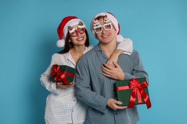 Noel Baba şapkalı ve pijamalı mutlu çift açık mavi arka planda Noel hediyeleri