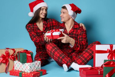 Noel Baba şapkalı ve pijamalı mutlu çift açık mavi arka planda Noel hediyeleri