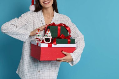 Noel Baba şapkalı ve pijamalı mutlu kadın. Açık mavi arka planda Noel hediyeleri.