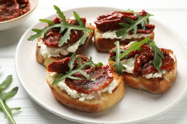 Krem peynirli lezzetli bruschettalar, güneşte kurutulmuş domatesler ve beyaz ahşap masada roka, yakın plan.