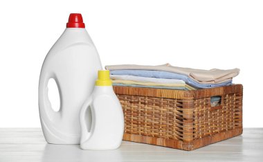 Temiz giysilerle çamaşır sepeti ve beyaz arka planda açık ahşap masada duran deterjan şişeleri.