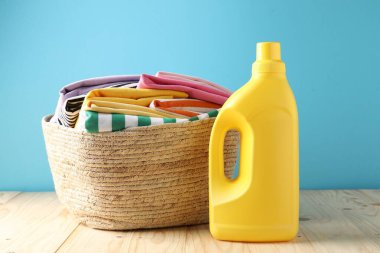 Açık mavi duvarın yanındaki ahşap masada çamaşır sepeti ve bir şişe deterjan.