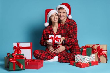 Noel Baba şapkalı ve pijamalı mutlu çift açık mavi arka planda Noel hediyeleri