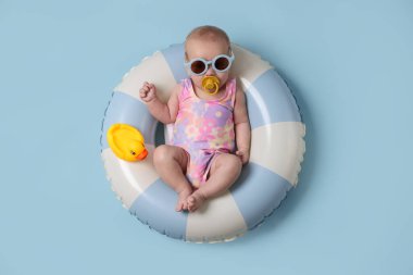 Güneş gözlüklü, oyuncak ördekli, şişme yüzüklü, açık mavi arka planda şirin bir bebek.