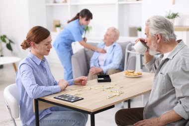 Masada yaşlı bir adamla domino oynayan ve hastasına huzur evinde yardım eden bir kadın.