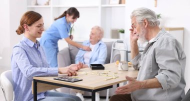 Masada yaşlı bir adamla domino oynayan ve hastasına huzur evinde yardım eden bir kadın.