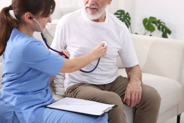 Bakım işçisi evdeki kanepede steteskopla yaşlı adamın kalp atışlarını kontrol ediyor.