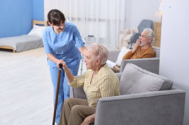 Bakım işçisi yaşlı kadına bastonuyla yardım ediyor huzurevinde koltuktan kalkması için.