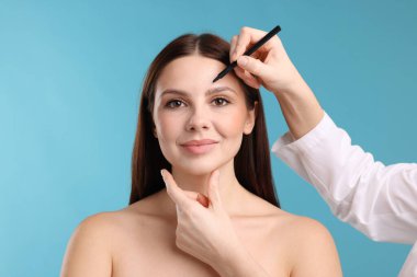 Doktor, açık mavi arkaplana karşı estetik ameliyat için hastanın yüzüne işaretler çiziyor.