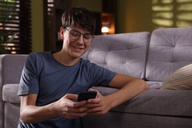 Evdeki kanepenin yanında akıllı telefon kullanan genç bir çocuk.