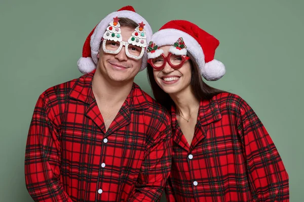 Noel Baba şapkalı mutlu çift, komik gözlükler ve gri yeşil arka planda pijamalar. Noel atmosferi