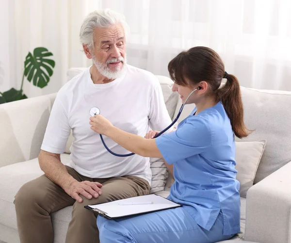 Bakım işçisi evdeki kanepede steteskopla yaşlı adamın kalp atışlarını kontrol ediyor.