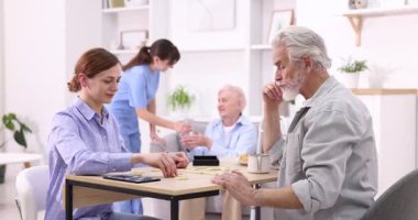 Masada yaşlı bir adamla domino oynayan ve hastasına huzur evinde yardım eden bir kadın.