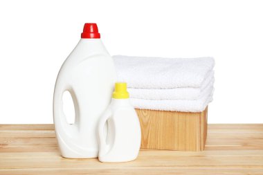 Çamaşır sepetinin içinde yumuşak havlular ve beyaz arka plandaki ahşap masada duran deterjan şişeleri.