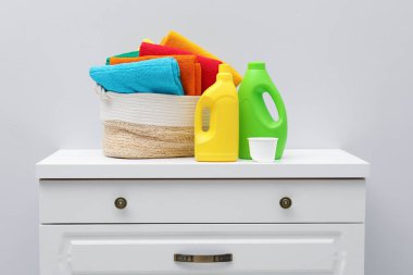 Çamaşır sepetinde havlular ve beyaz duvarın yanındaki dolapta deterjan var.