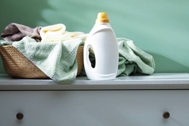 İçerideki yeşil duvarın yanındaki şifonyerin üstünde havlu ve bir şişe deterjan olan çamaşır sepeti.