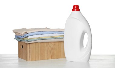 Temiz elbiseli çamaşır sepeti ve beyaz arka planda açık ahşap masa üzerinde bir şişe deterjan.