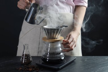 Kadın, siyah masadaki filtreli kimya kahve makinesine sıcak su döküyor. Siyah arka planda dumanlı, yakın plan.