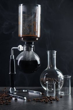 Vakumlu kahve makinesinde aromatik kahve demlemek, laboratuvar bardağı ve fasulye gri masada koyu arkaplanda dumanla kaplanmak, yakın plan