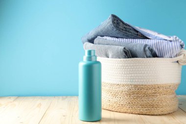 Açık mavi duvarın yanındaki ahşap masada çamaşır sepeti ve bir şişe deterjan.