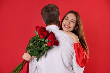 Kırmızı arka planda elinde gül buketiyle erkek arkadaşına sarılan mutlu bir kadın. Sevgililer Günü kutlaması