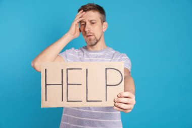 Unhappy man holding cardboard sign with word Help on light blue background
