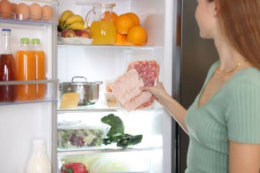 Mutfaktaki buzdolabının rafına jambon ve salam koyan kadın, yakın plan.