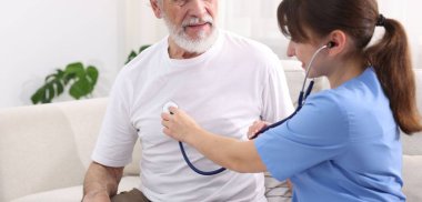 Bakım işçisi evdeki kanepede steteskopla yaşlı adamın kalp atışlarını kontrol ediyor.