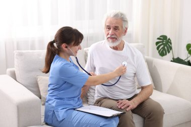 Bakım işçisi evdeki kanepede steteskopla yaşlı adamın kalp atışlarını kontrol ediyor.