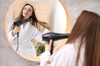 Bornozlu güzel bir kadın banyoda aynanın yanında saç kurutma makinesiyle saçını kurutuyor.