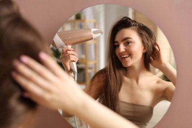 Gülümseyen kadın saçını kurutan saç kurutma makinesiyle içeride aynanın yanında.