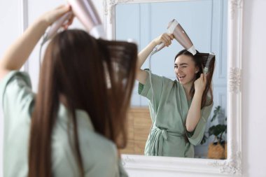 Bornozlu mutlu kadın saçını saç kurutma makinesiyle şekillendiriyor ve aynanın yanında kapalı bir yerde tarıyor.