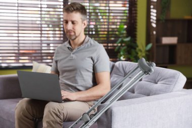 Evdeki koltukta dizüstü bilgisayar kullanan adam koltuk değneklerine odaklan.