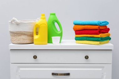 Çamaşır sepetinde havlular ve beyaz duvarın yanındaki dolapta deterjan var.