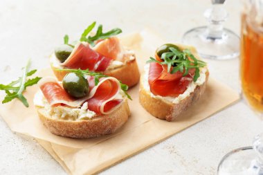 Krem peynirli lezzetli bruschettalar, prosciutto, yeşil zeytin ve roka gri masada, yakın plan.