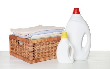 Temiz giysilerle çamaşır sepeti ve beyaz arka planda açık ahşap masada duran deterjan şişeleri.