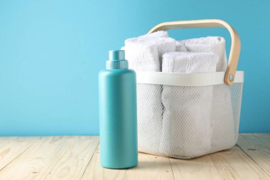 Açık mavi duvarın yanındaki ahşap masanın üzerinde havlu ve bir şişe deterjan olan çamaşır sepeti.