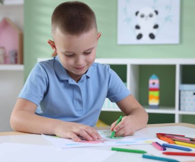Cute little boy drawing with crayon at table indoors