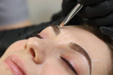 Güzellik uzmanı, kapalı mekanda kalıcı kaş makyajı prosedüründen önce kadının yüzüne işaretler çiziyor.