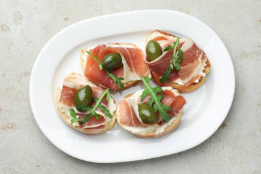 Krem peynirli nefis bruschettalar, prosciutto, yeşil zeytin ve roka, gri desenli masa, üst görünüm